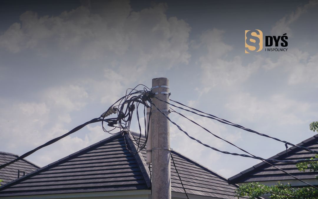 Przyłącze kablowe na słupie przed dachami budynków w pasie technologicznym linii energetycznej.